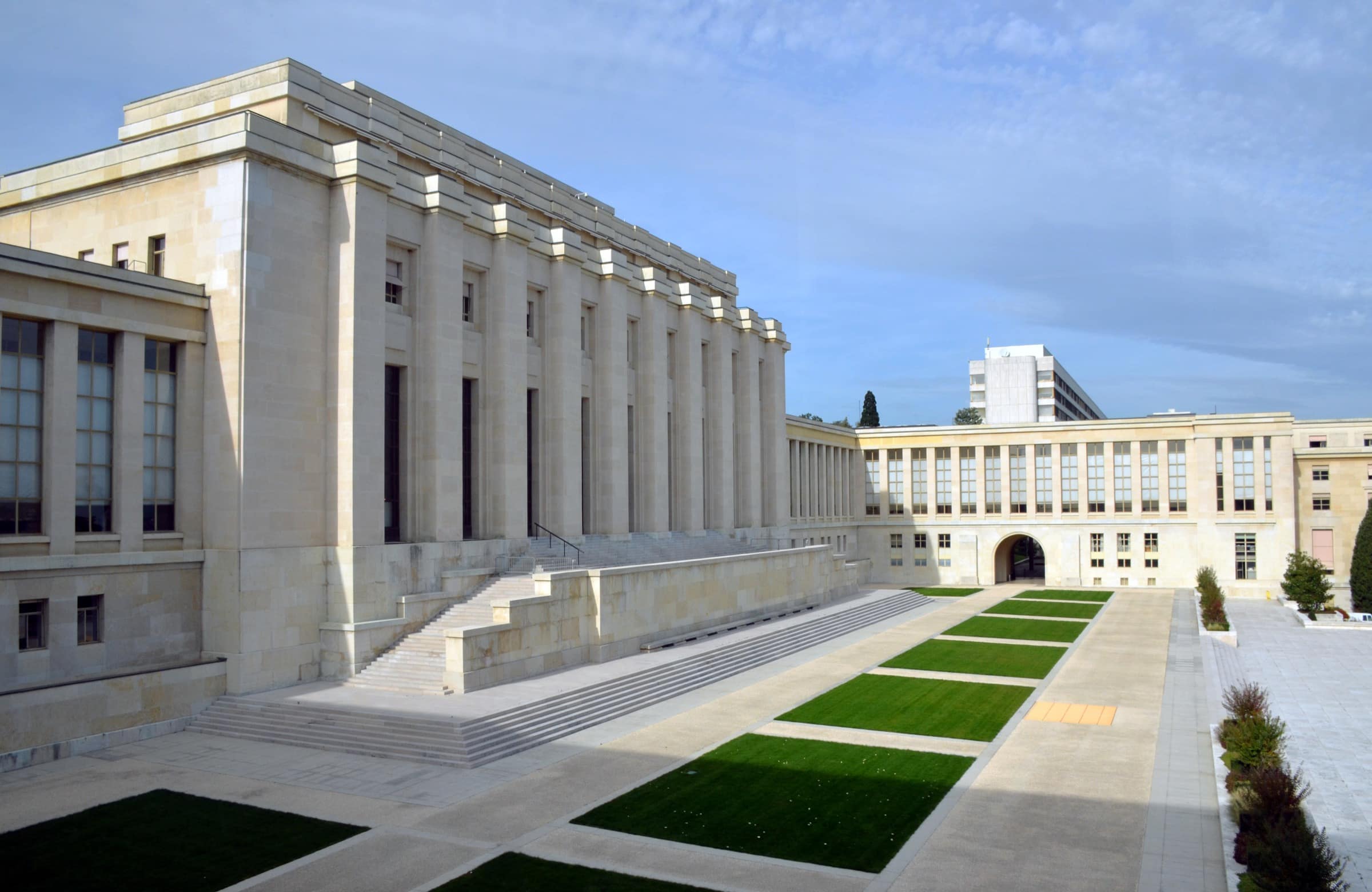 The Palais des Nations in Geneva seen across its Cour d'Honneur: a long pale stone facade with a deep colonnade and a broad flight of steps at left, a lower glazed wing closing the courtyard ahead, and rectangles of lawn set into the paving.