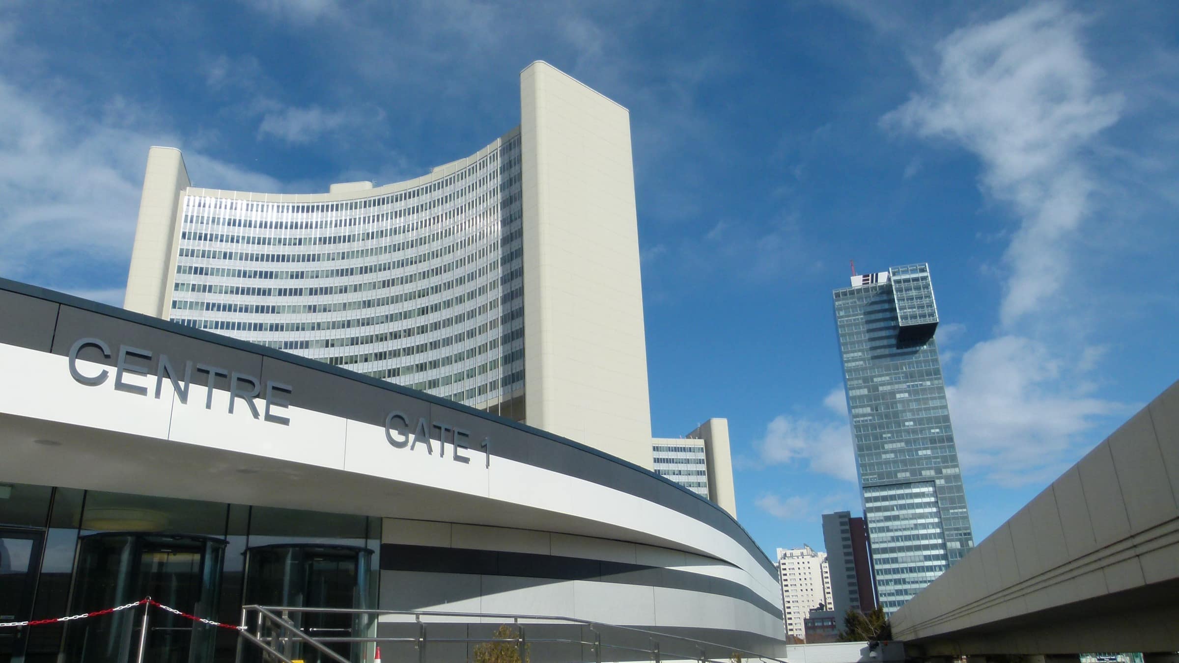 The curved visitor-entrance canopy of the Vienna International Centre, lettered CENTRE GATE 1 in plain capitals along its fascia, with one of the complex’s curved office towers rising behind it against an open sky.