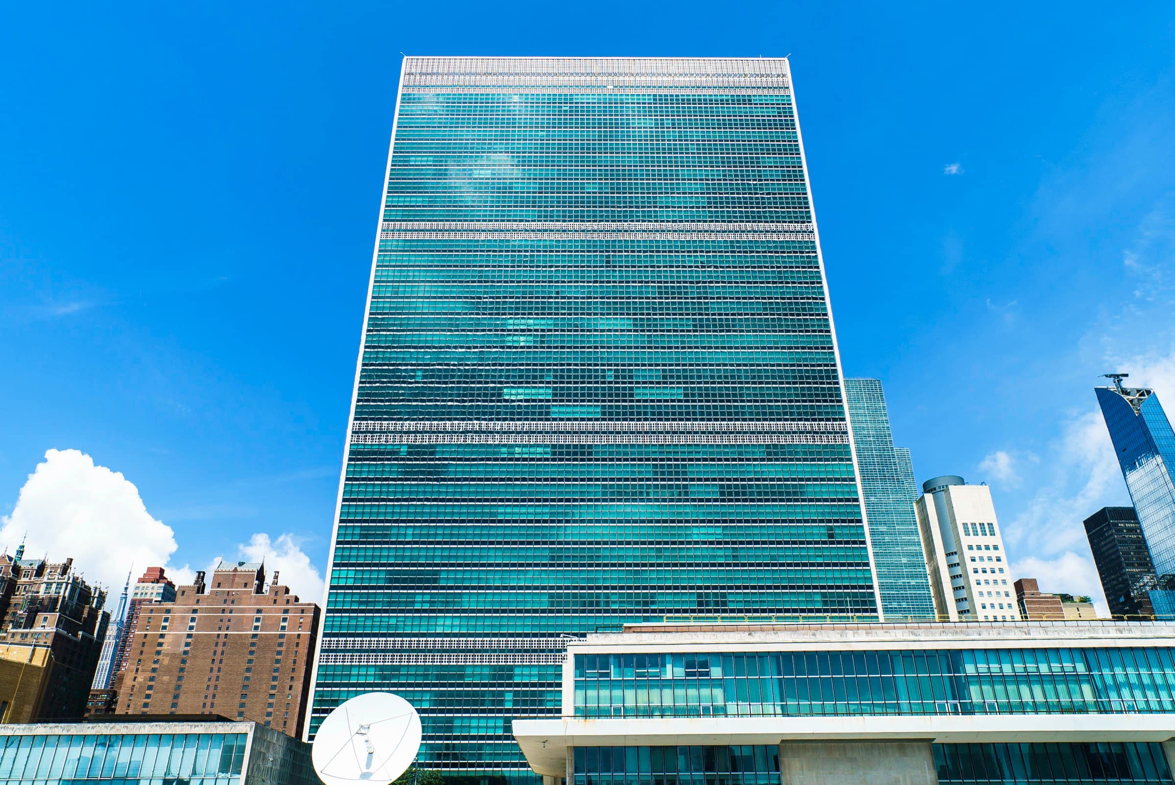 The United Nations Secretariat tower in New York seen from below against a clear blue sky, its glass curtain wall banded by service floors, with the low roof of the Conference Building and a satellite dish in the foreground and midtown towers at either side.